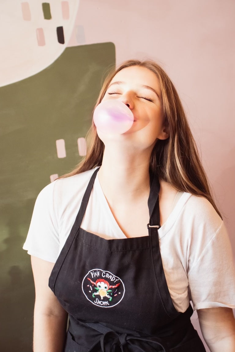 A you woman with long blonde hair has her eyes closed and her head back blowing a big pink bubble gum bubble.